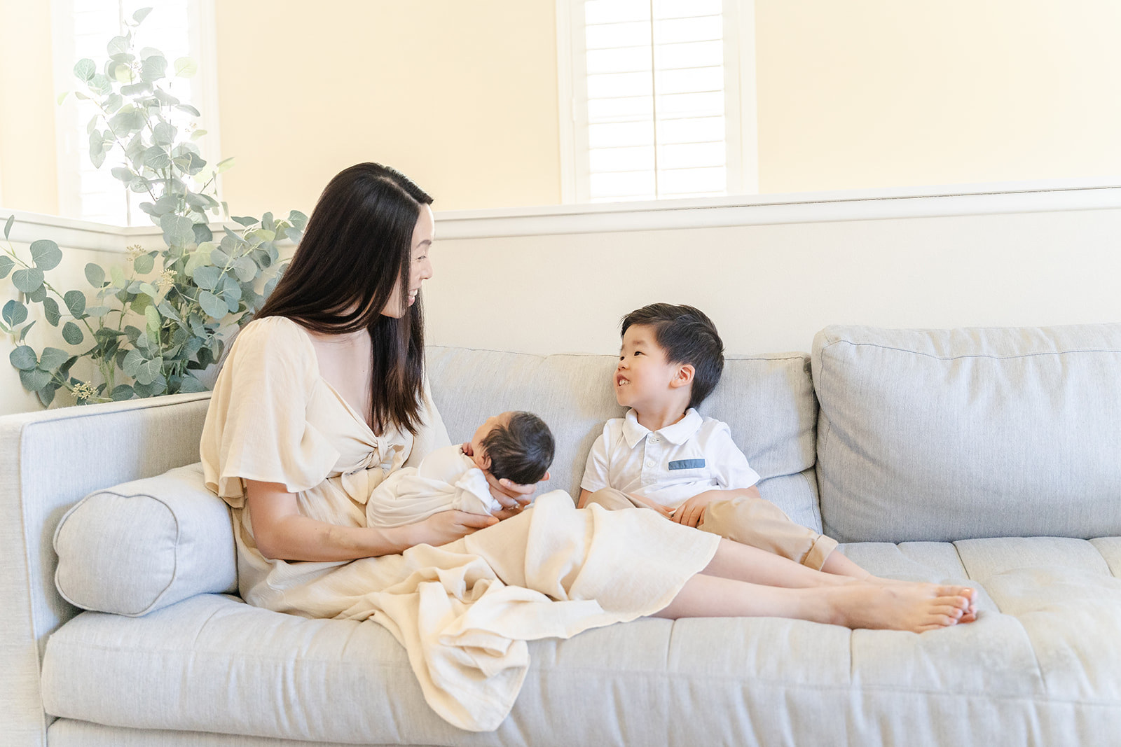 A mother in a tan dress sits across a couch with her newborn baby in her lap and toddler son next to her