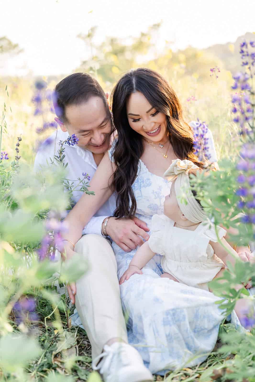 A happy mom and dad snuggle in purple wildflowers with their infant in their lap