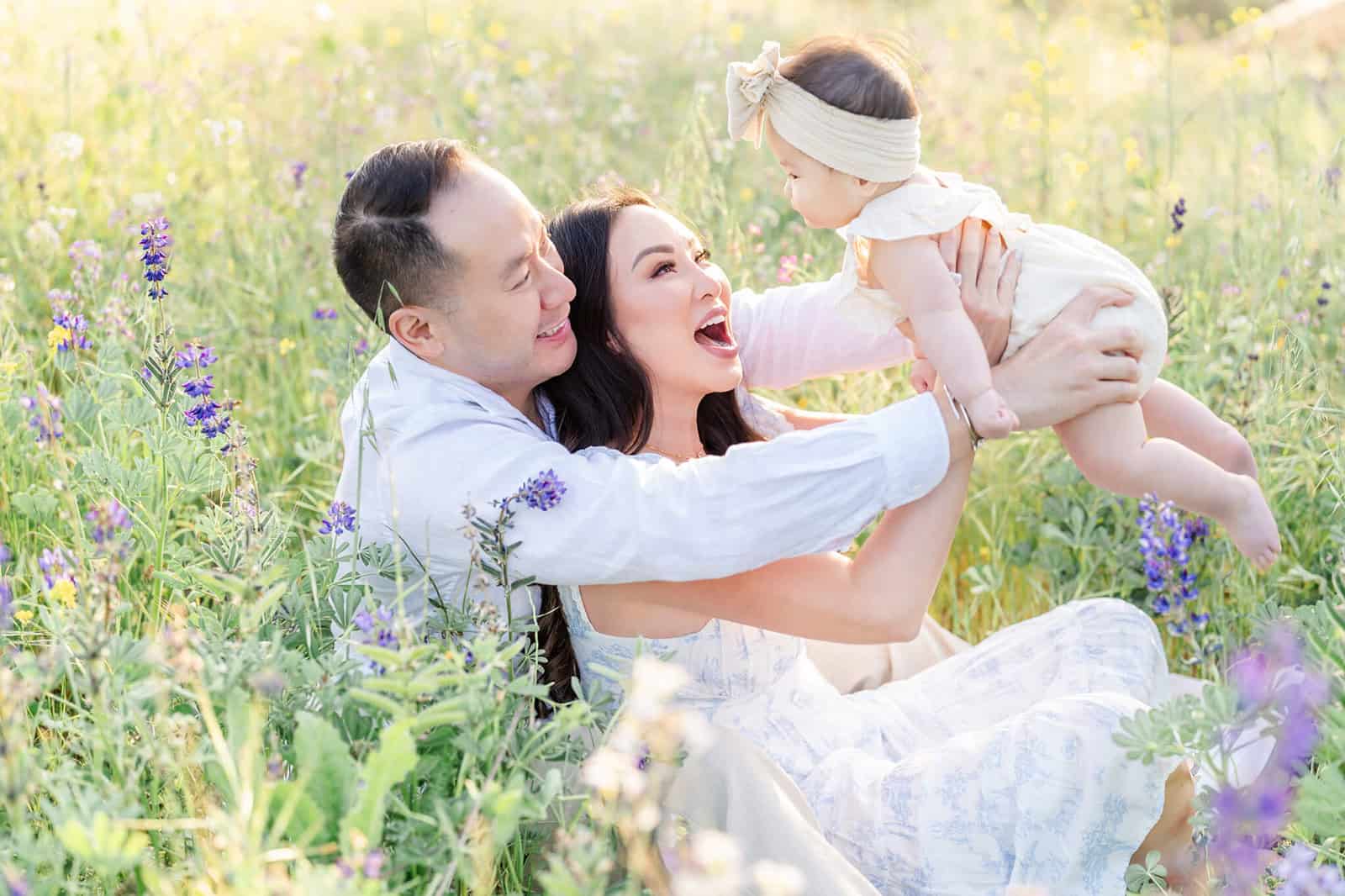Happy mom and dad sit among purple wildflowers lifting and playing with their baby girl at sunset