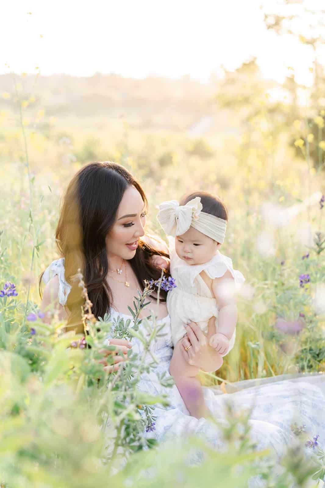 A happy mother in a white dress plays in wildflowers with her infant daughter at sunset after visiting Kidspace Children's Museum