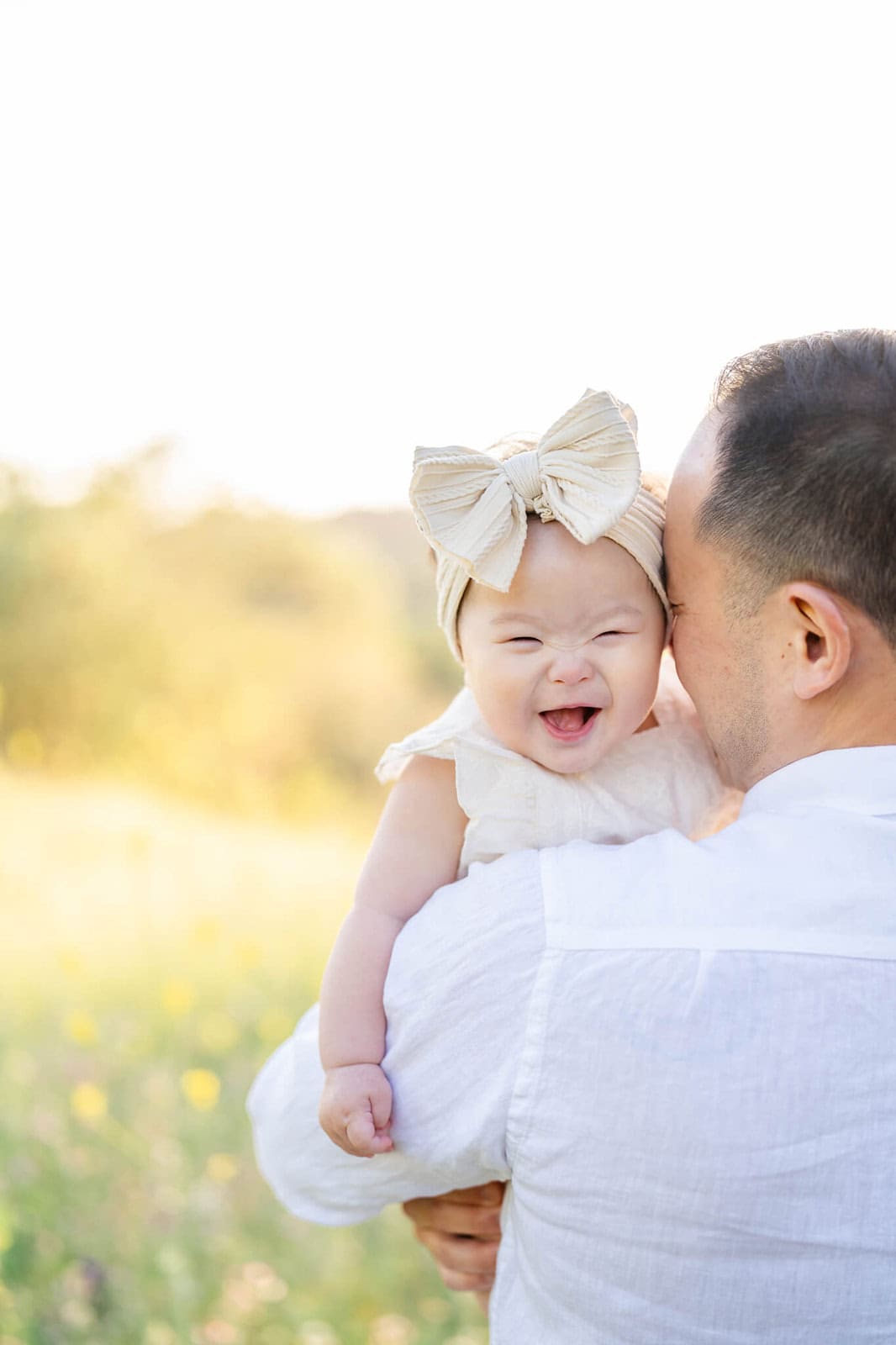 A giggling baby on dad's shoulder with a big bow headband at sunset in a meadow after visiting Kidspace Children's Museum