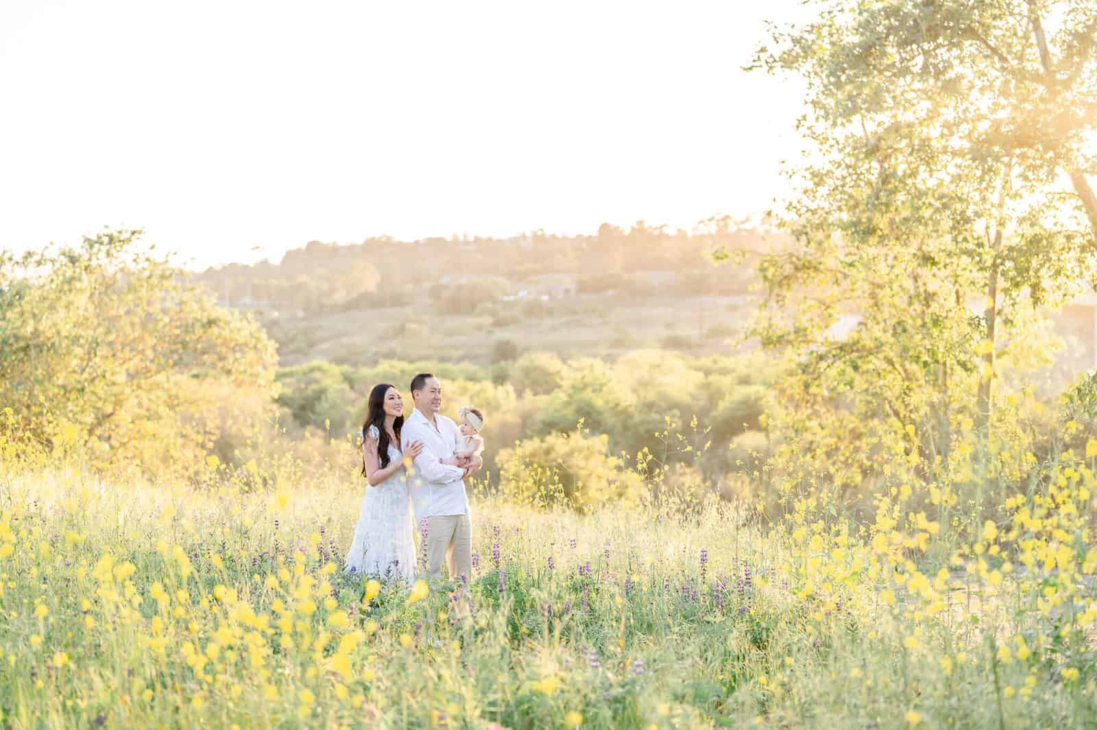 A mom and dad stand on a flower covered hillside at sunset holding their baby girl before visiting Kidspace Children's Museum