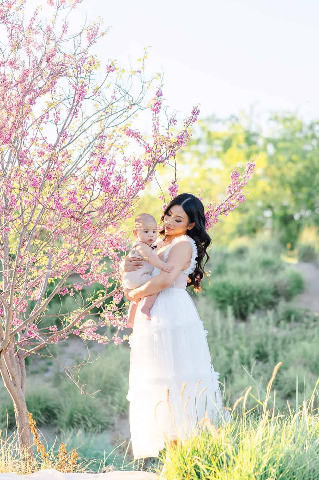 A woman stands holding her infant in her arms against her chest under a young pink blooming tree at sunset