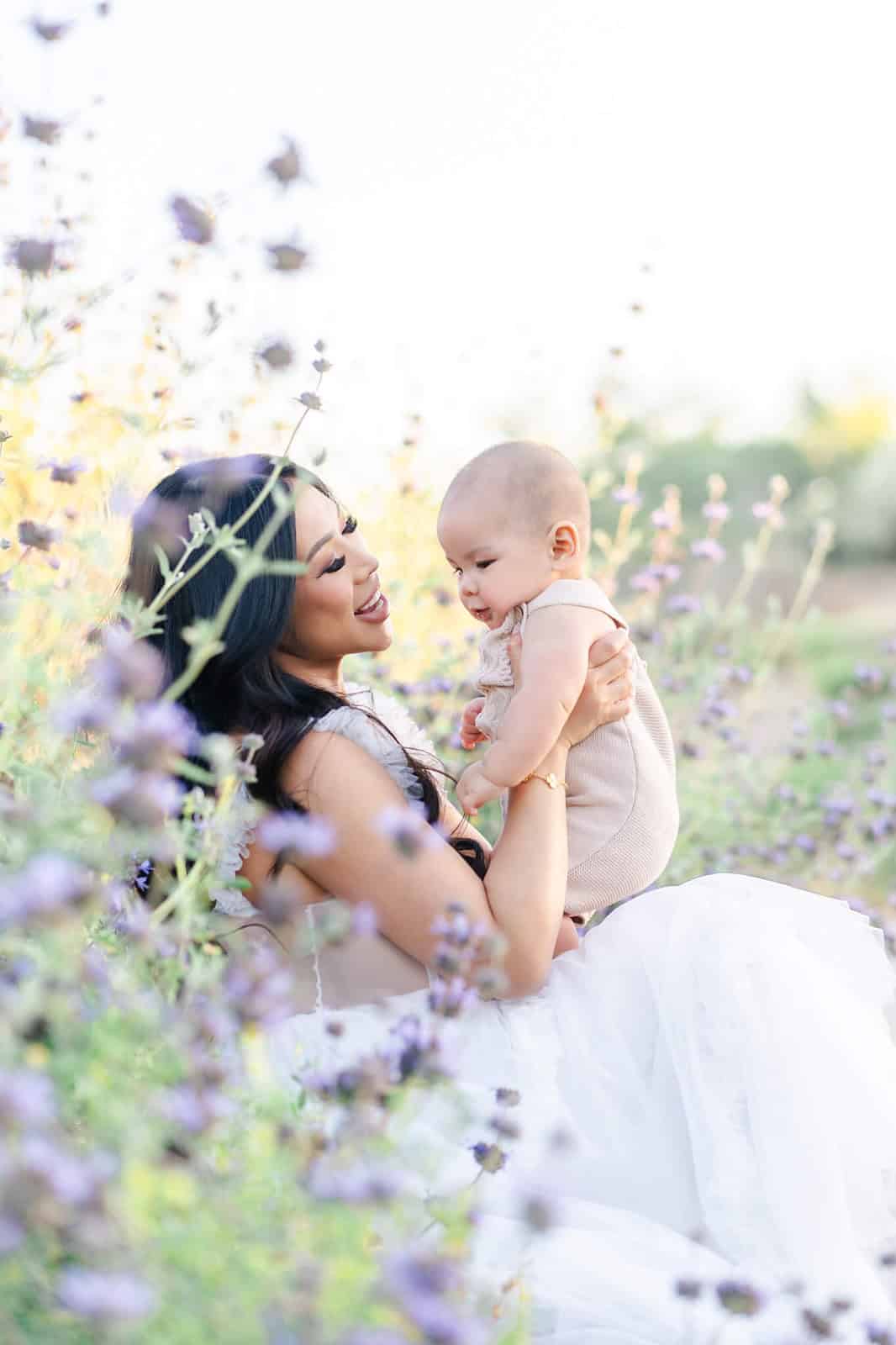 A mother sits in purple flowers in a garden wearing a white gown playing with her infant in her lap during things to do in LA with toddlers