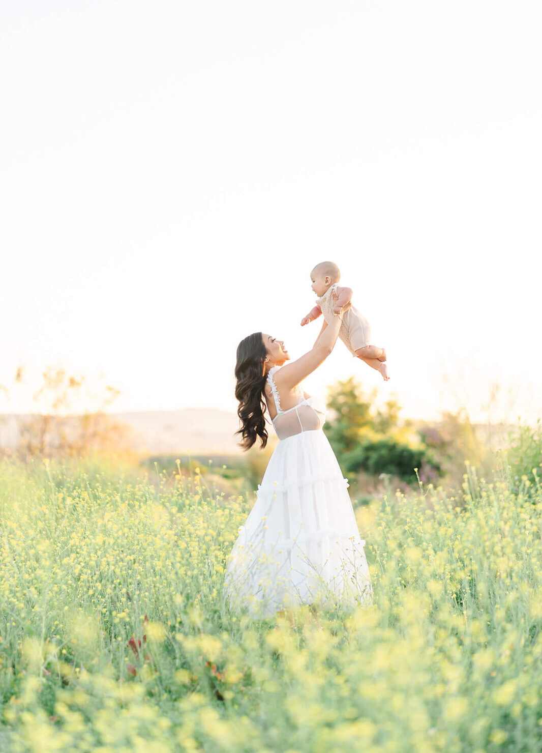 A happy new mother lifts and plays with her infant in a field of yellow wildflowers at sunset after finding things to do in LA with toddlers