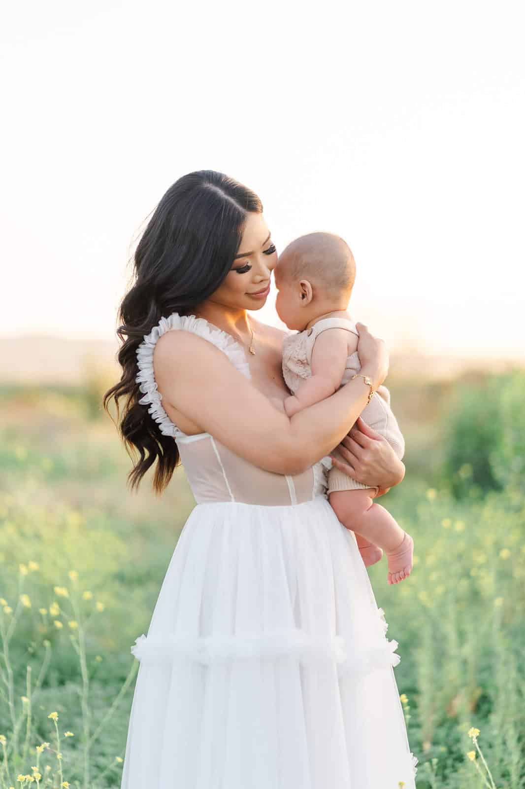 A new mom in a white gown stands in a field of wildflowers at sunset holding her newborn after finding things to do in LA with toddlers