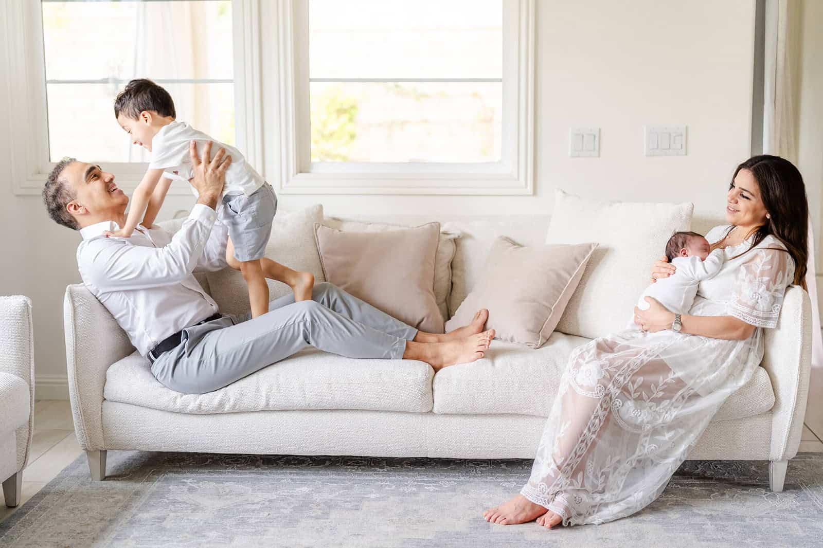 A dad plays with his toddler son on a couch with mom cradling newborn on her chest on the other side of the couch