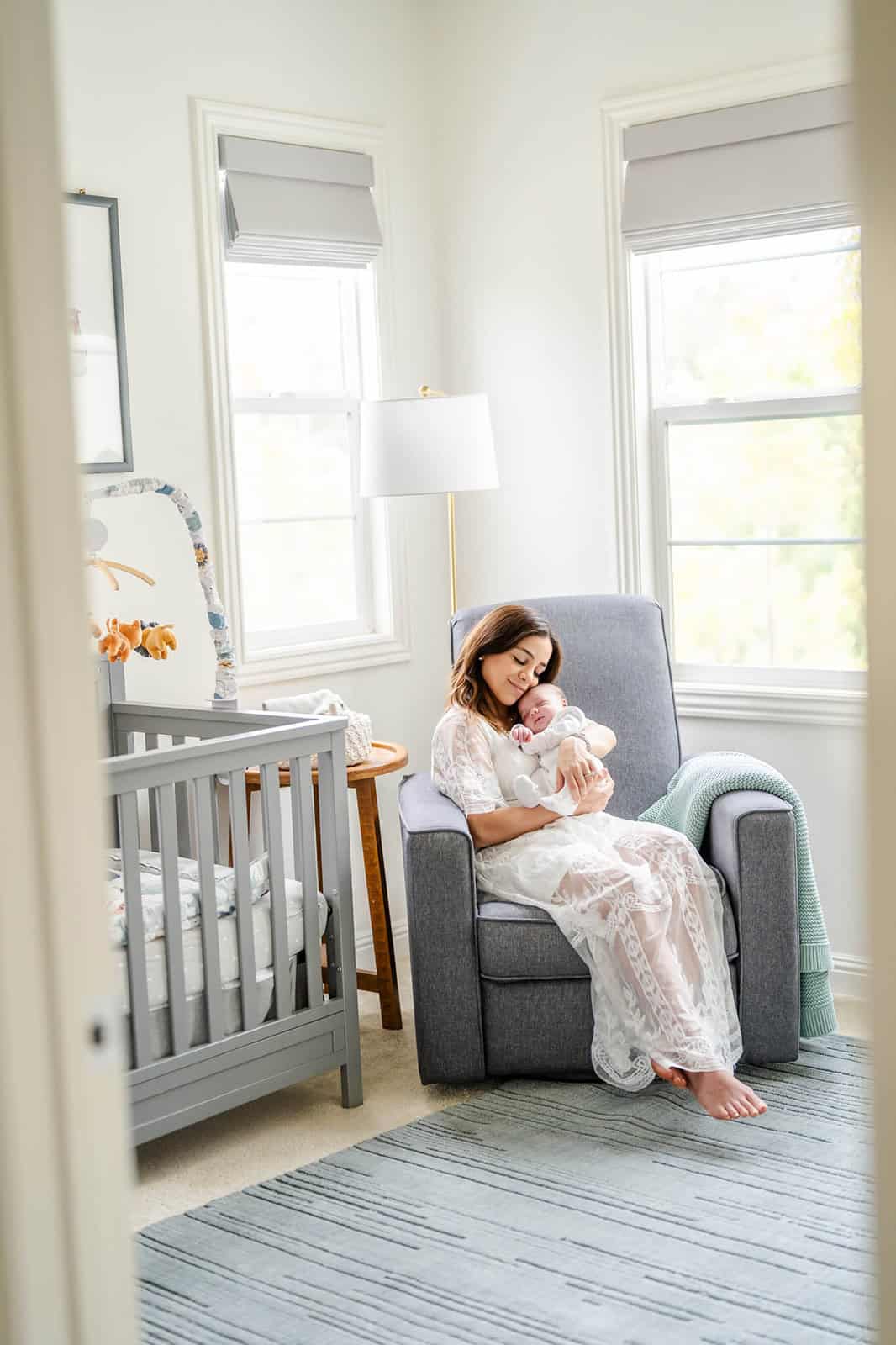 A happy mother in a white lace gown sits in a nursing chair under windows cradling her newborn against her cheek