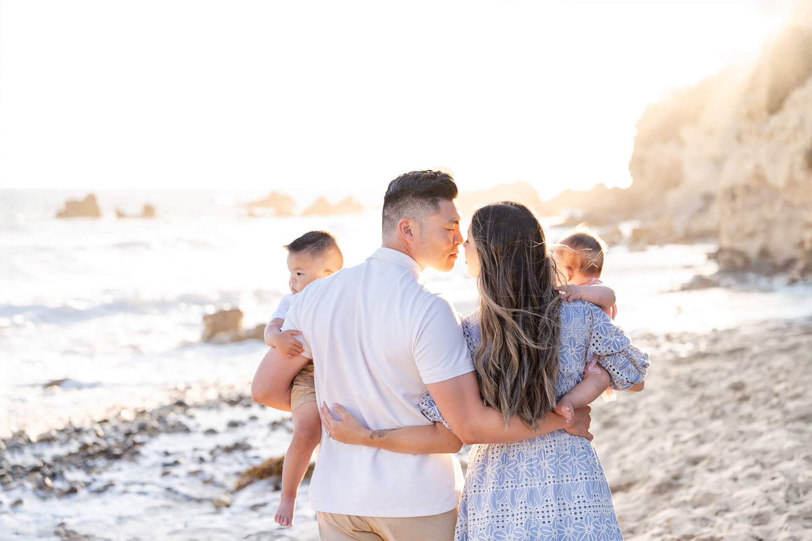 Happy mom and dad lean in for a kiss while walking on a beach at sunset carrying their toddler son and daughter