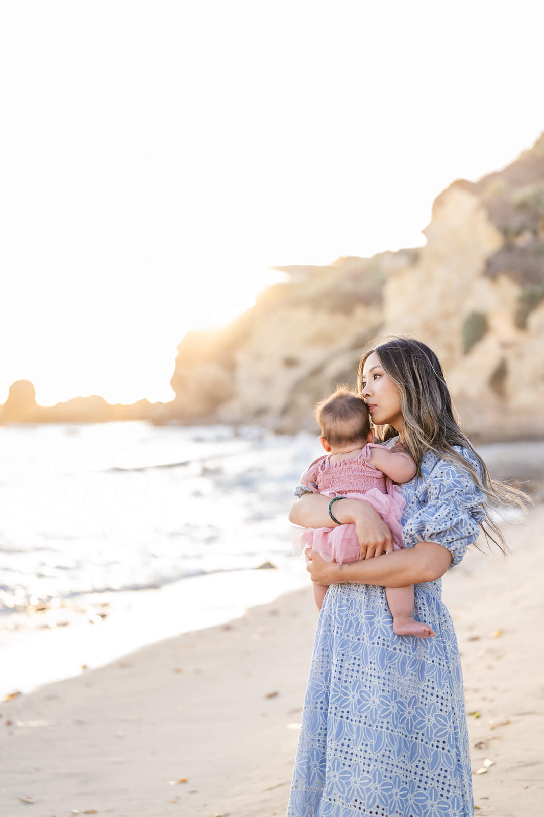 A mother in a blue dress snuggles her toddler daughter in pink on a beach at sunset after visiting Marina Park Newport Beach