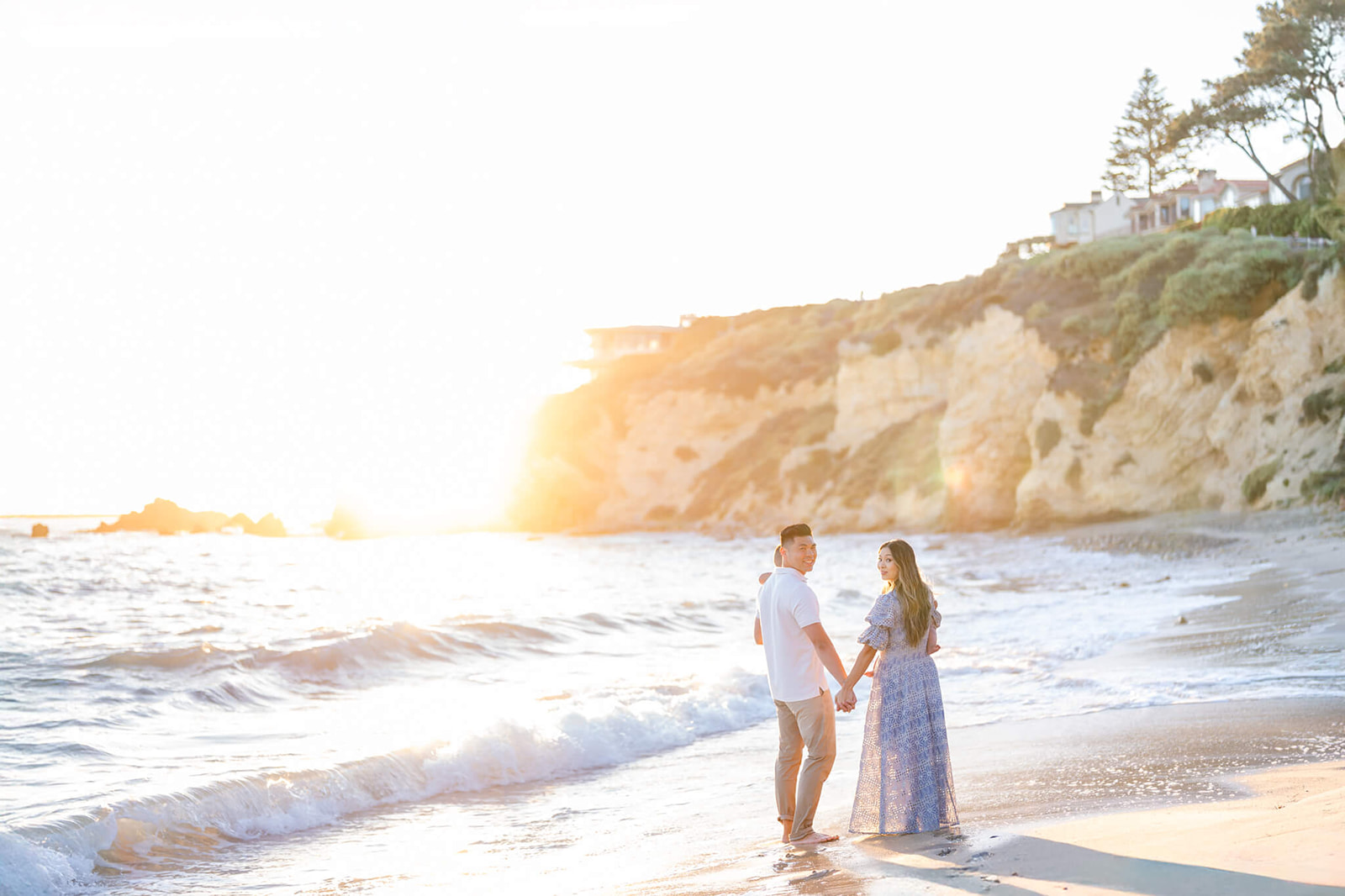 A smiling couple hold hands in blue and white while walking on a beach holding their two toddlers before visiting Marina Park Newport Beach