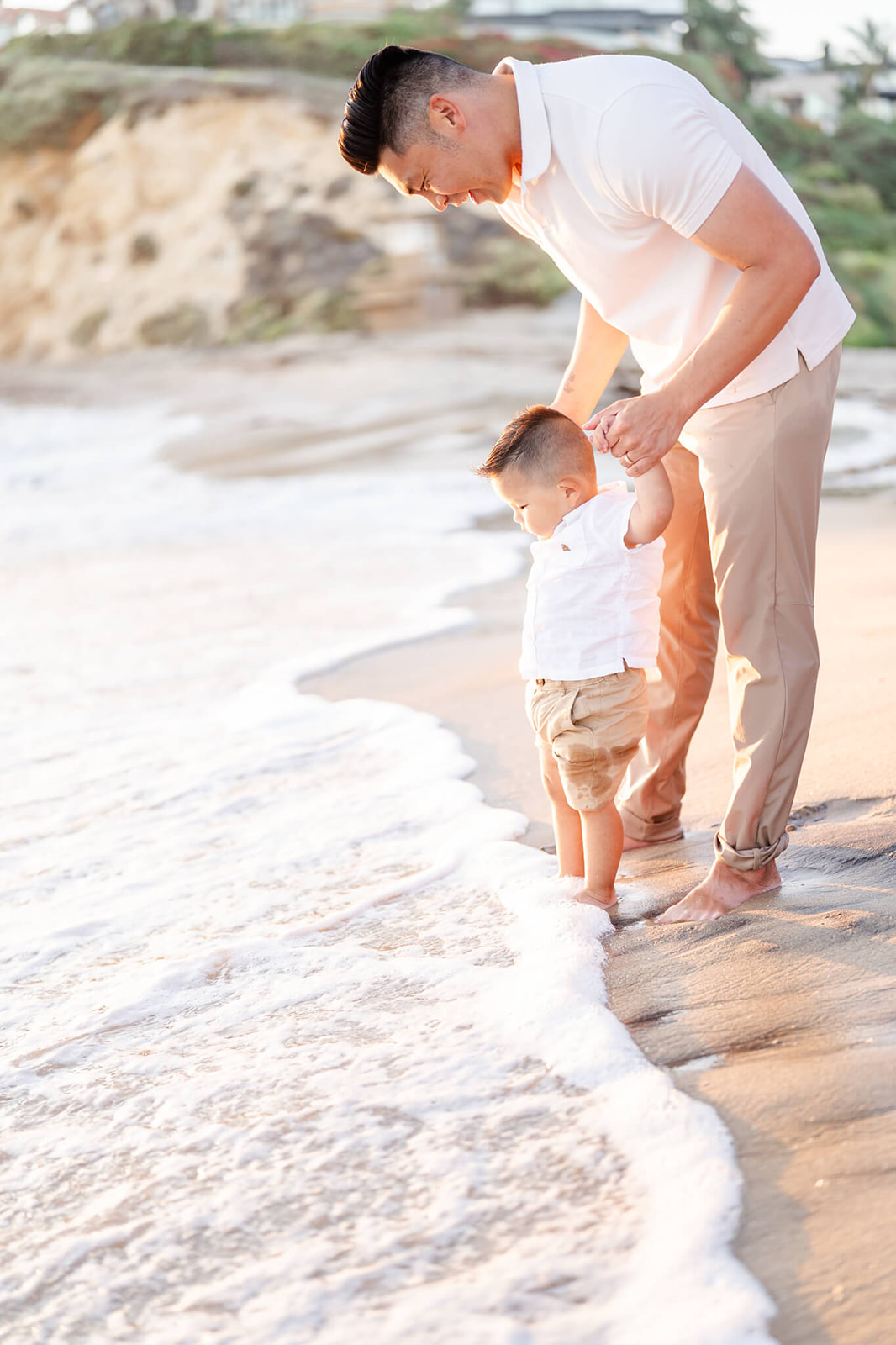 A happy dad in white and khakis helps his son feel the water on his beach on a beach when visiting Marina Park Newport Beach