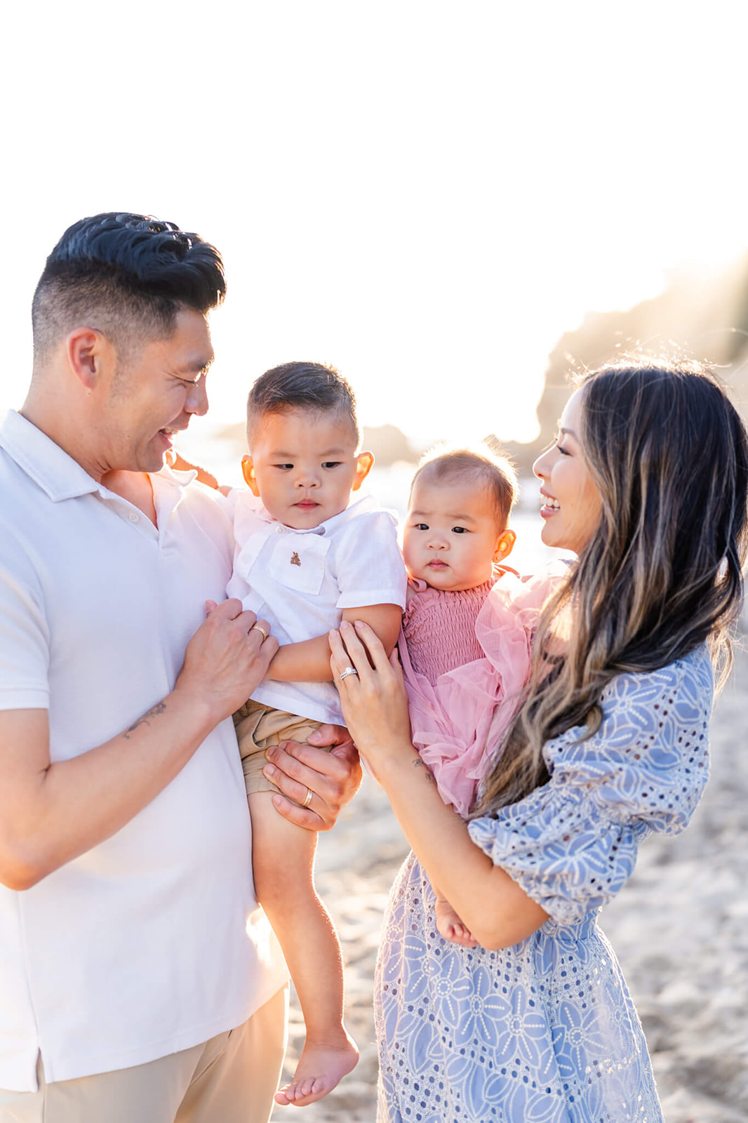 Happy mom and dad laugh while standing on a beach holding their toddler son and daughter between them