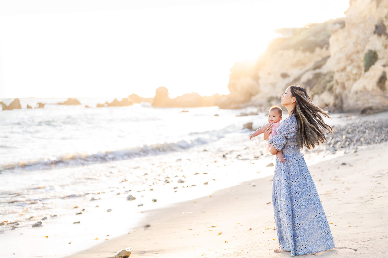 A mom stands in a blue gown holding her toddler daughter in pink on a beach at sunset