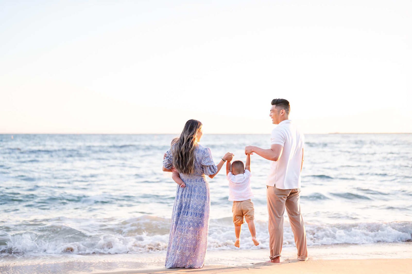 A mom and dad lift their toddler by the hands at sunset on a beach in white and blue before visiting Seascape Kids Fun