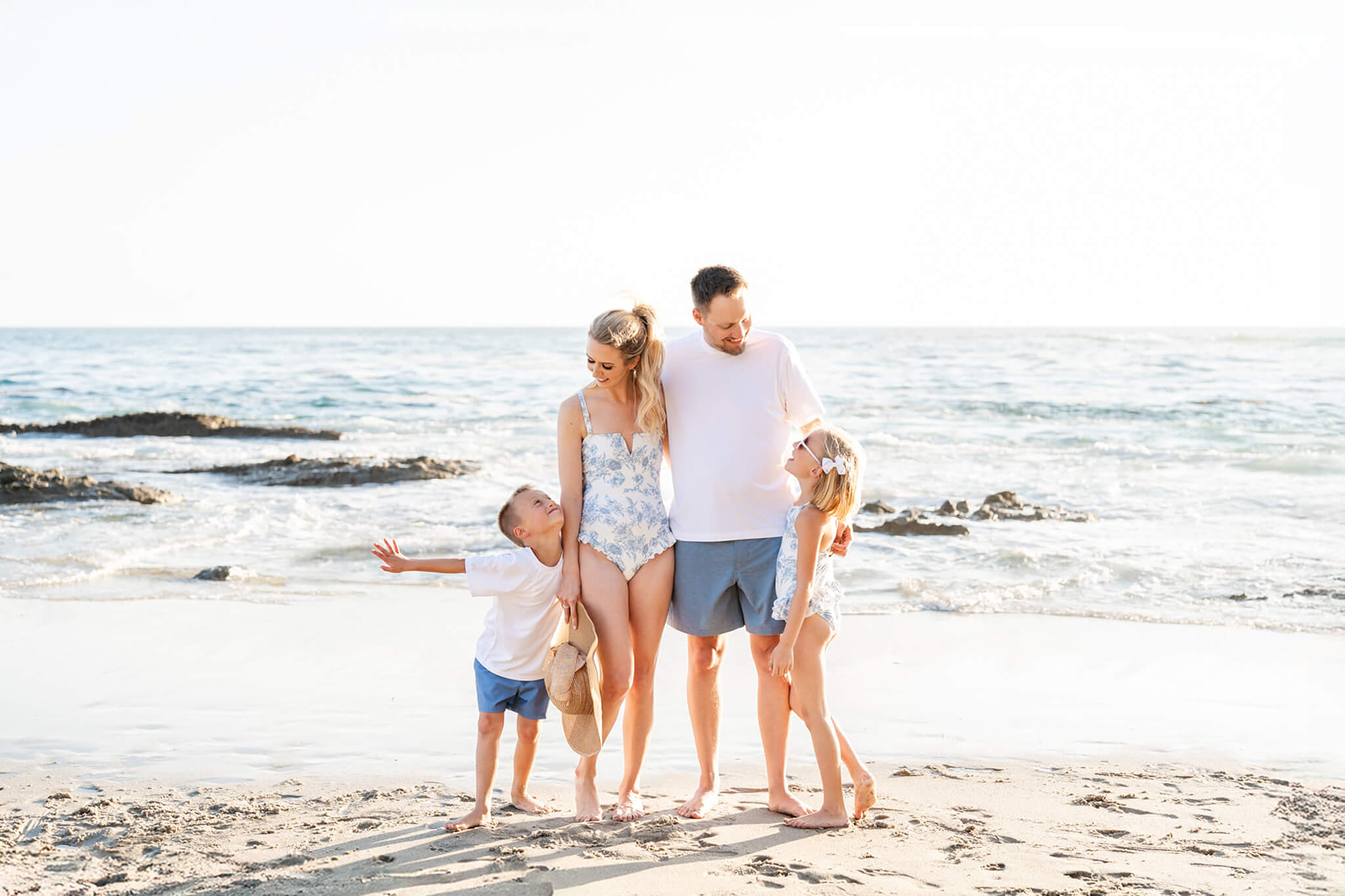 A happy mom and dad stand on a beach smiling down to their toddler son and daughter in matching blue and white bathing suits after visiting Wild Rivers Irvine