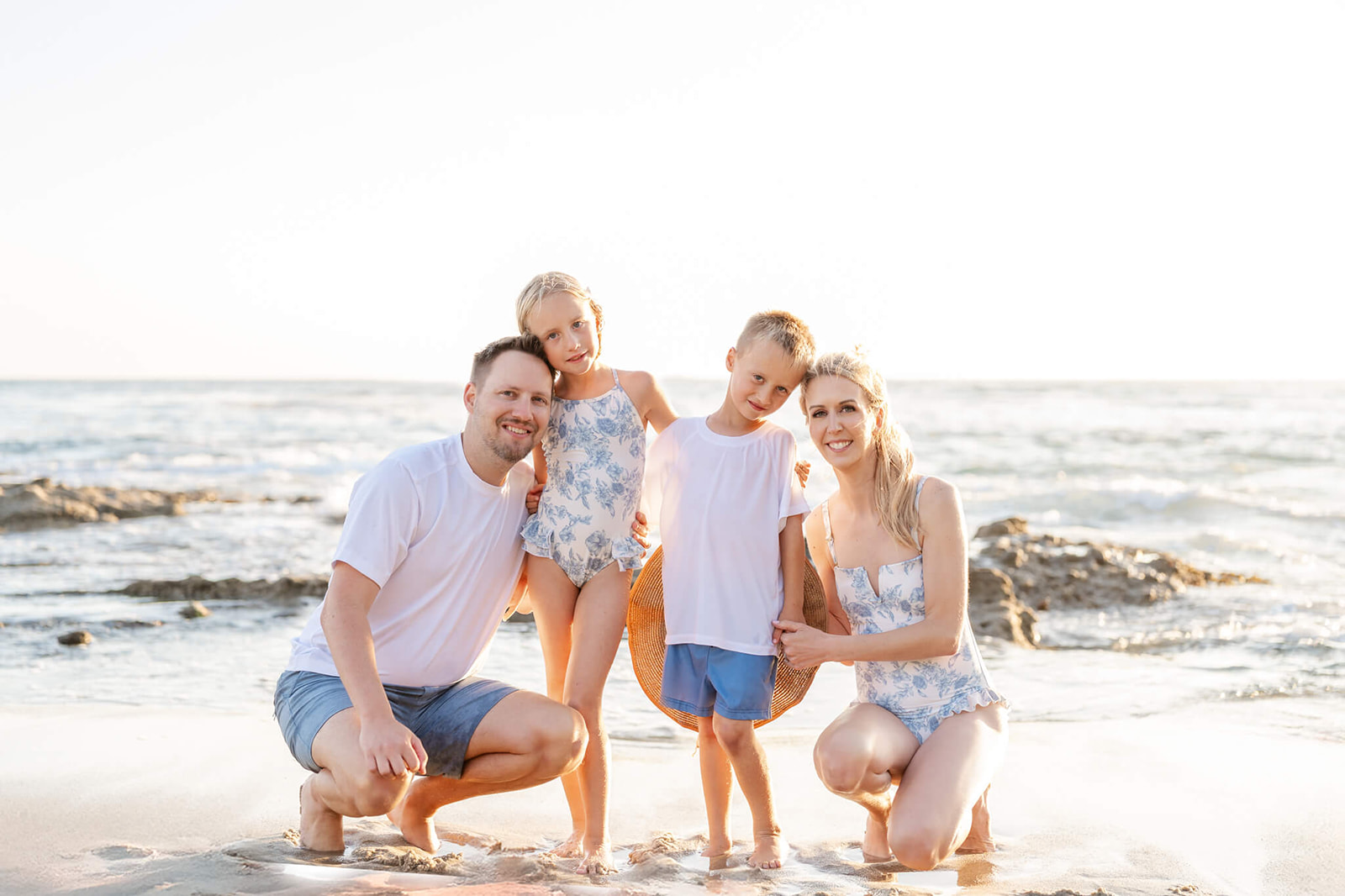 A mom and dad on a beach in blue and white hug and smile with their toddler son and daughter in matching bathing suits before heading to Wild Rivers Irvine