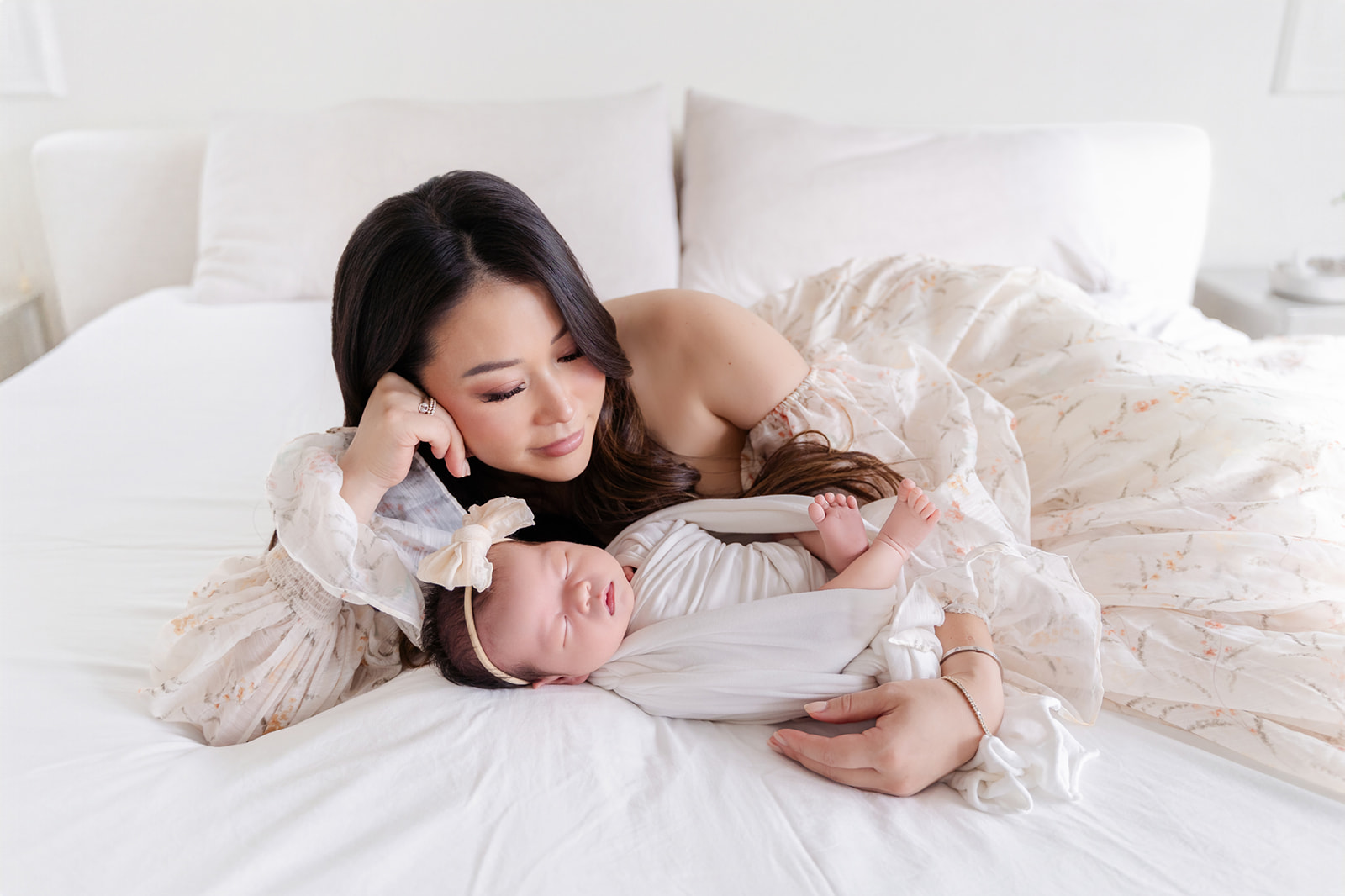 A happy new mom lays across a bed in a floral print gown cradling her newborn daughter