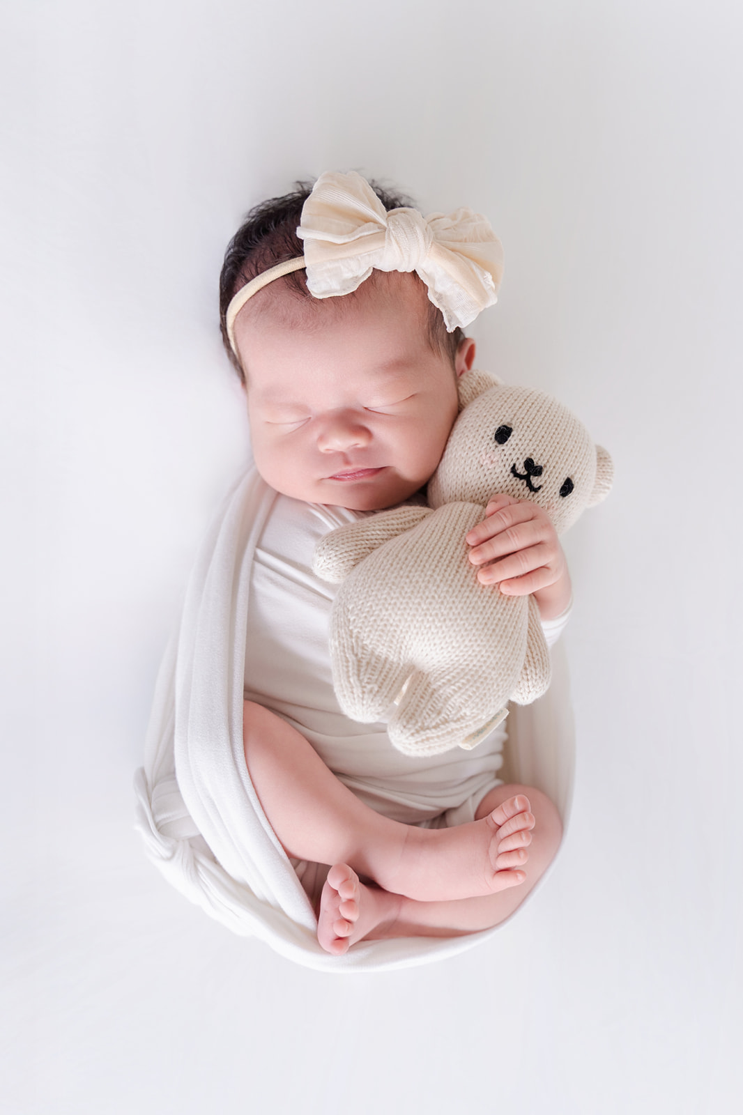 A sleeping newborn baby cuddling a stuffed bear in a tiny bow in a white swaddle after finding Irvine Chinese Immersion Academy