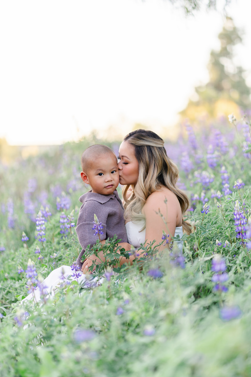 A mother kisses the cheek of her toddler son in her lap in a grey sweater sitting in a park full of wildflowers