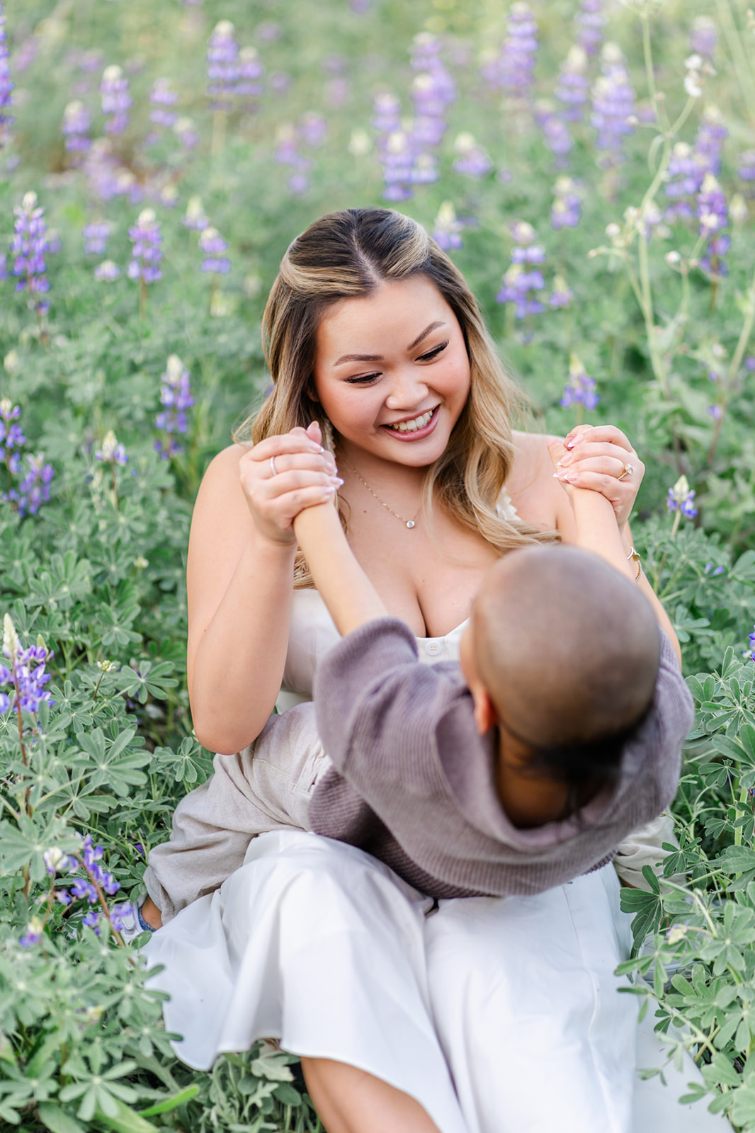 A mom plays with her toddler son in her lap in a white dress while sitting among purple wildflowers at sunset while visiting Irvine parks