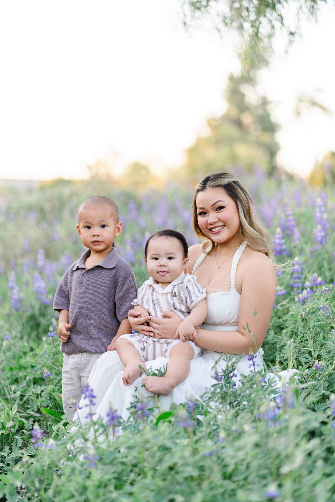 A happy mom in a white dress sits in a field of wildflowers at sunset with her two very young children in her lap at one of the Irvine parks
