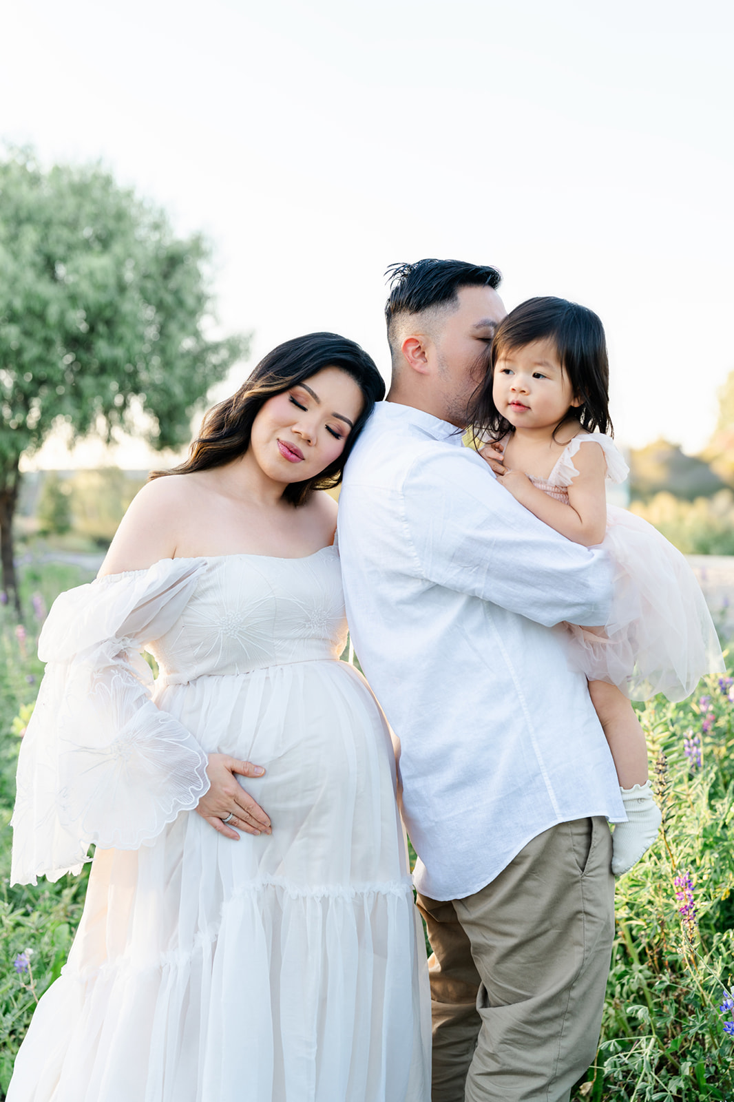 A pregnant mom in a white maternity gown snuggles onto the shoulder of her husband in white holding their toddler daughter