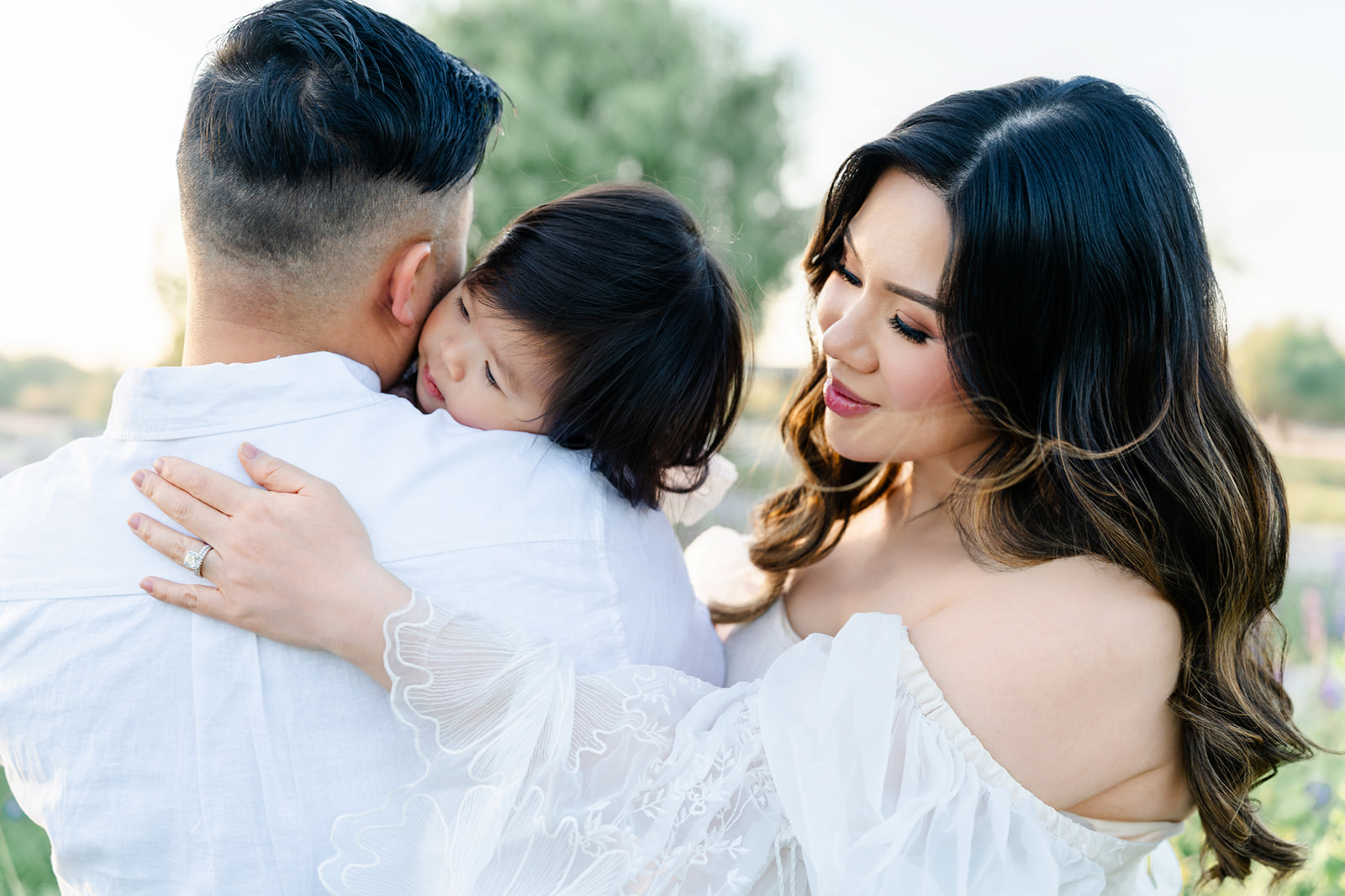 A happy mom and dad snuggle their toddler daughter on dad's shoulder at sunset in white