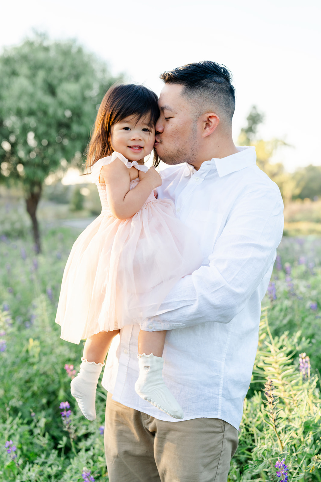 Dad snuggles and kisses the cheek of his happy toddler daughter while standing in a field of purple wildflowers
