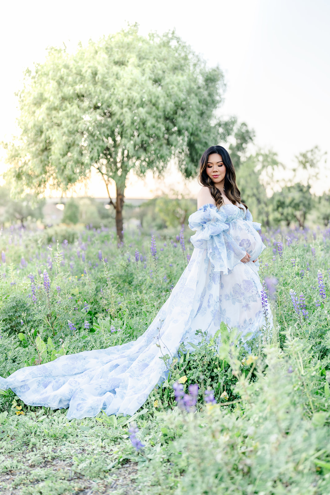 A mother to be in a long flowing blue maternity gown walks through purple wildflowers at sunset before visiting Candeeland Santa Ana