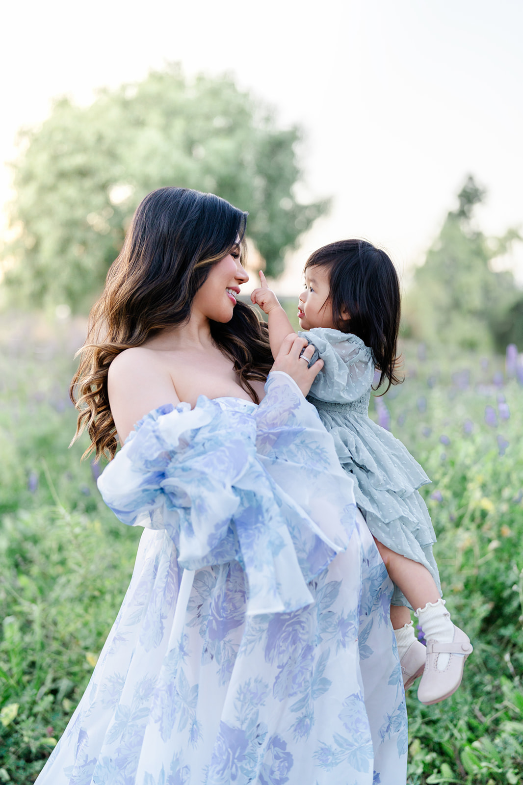 A happy mom plays with her toddler daughter on her bump in a garden in a blue maternity gown before visiting Candeeland Santa Ana