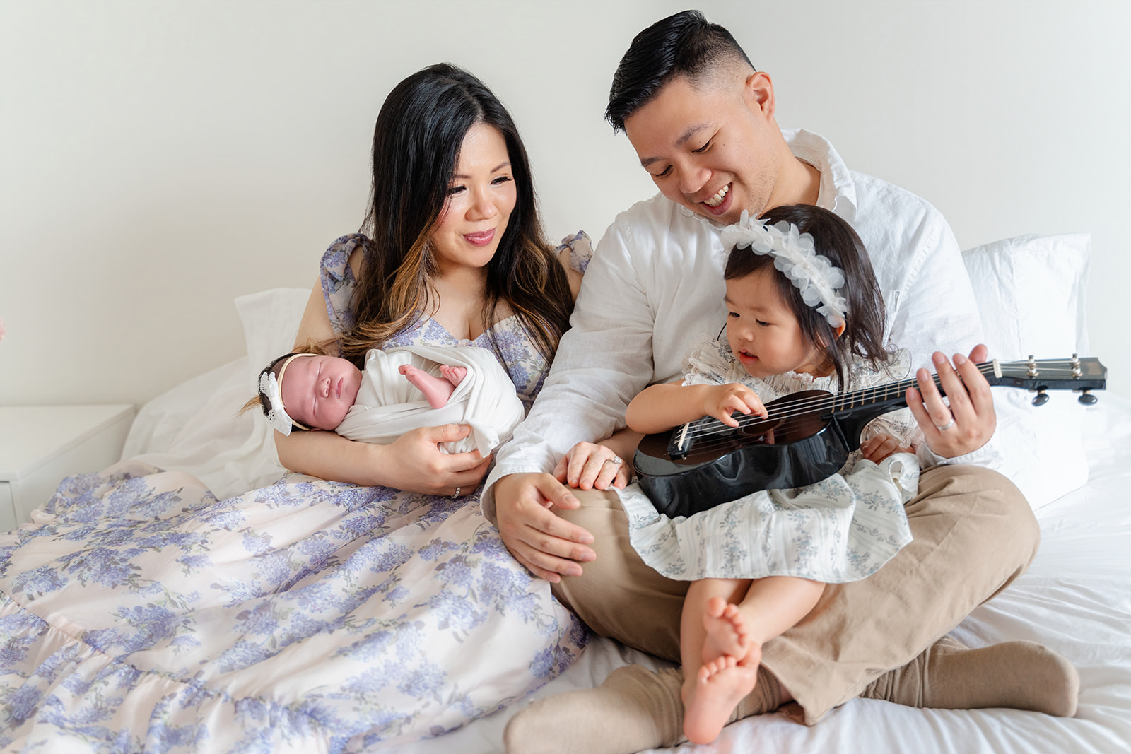 A toddler girl strums a ukelele while sitting in dad's lap next to mom holding newborn