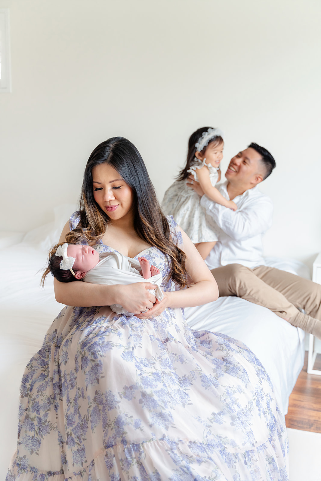 A mother cradles her sleeping newborn in her arms while dad plays with toddler on the bed behind her