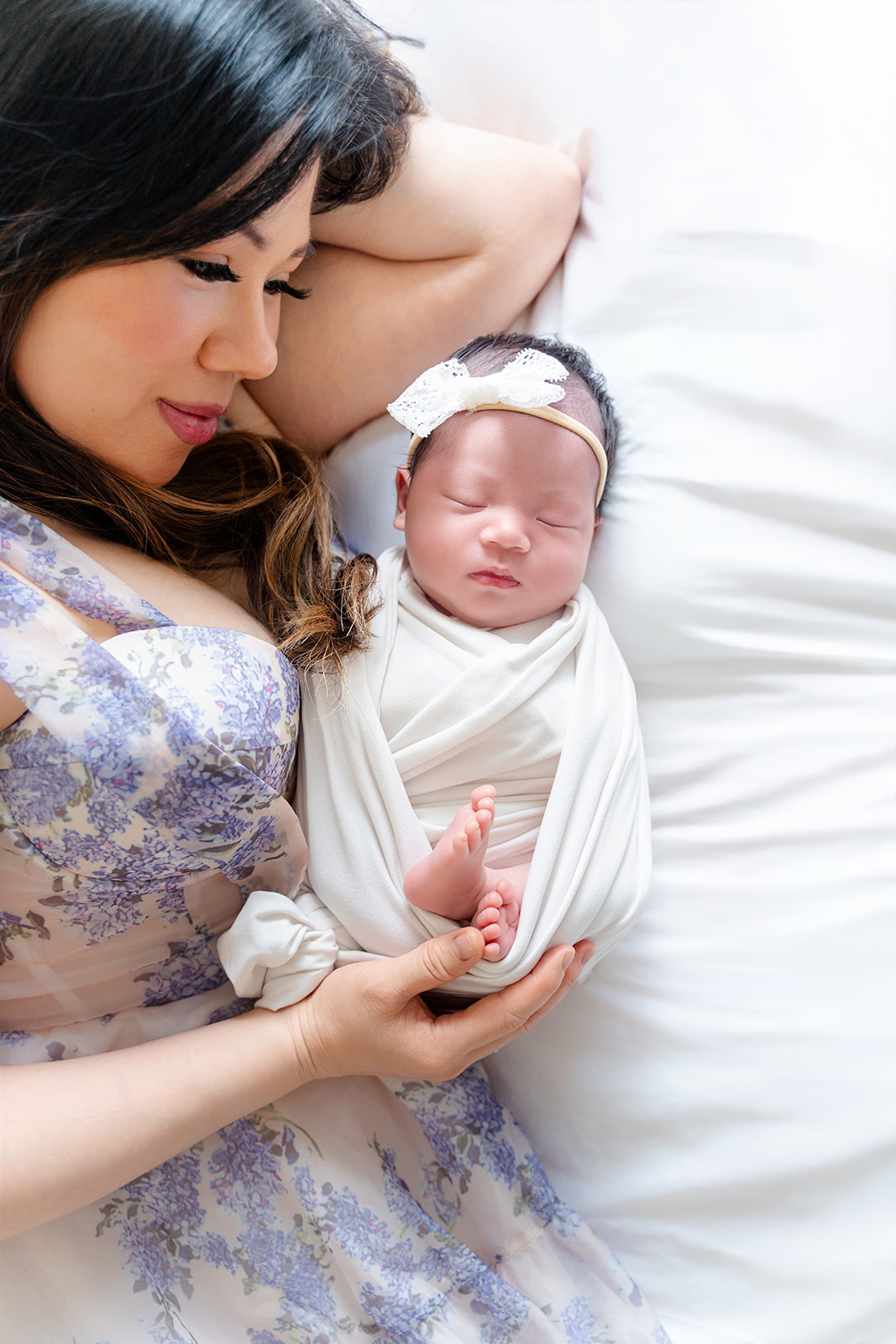 A new mom in a blue dress snuggles her sleeping newborn daughter in a white swaddle and bow headband