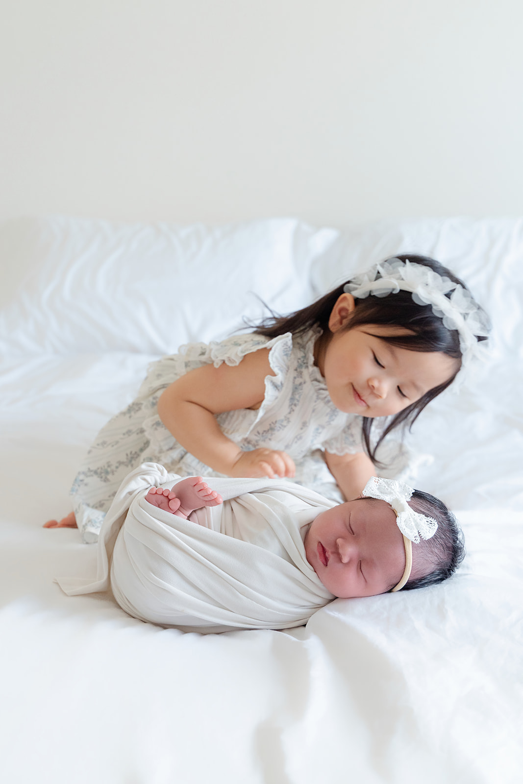 A toddler girl on a bed reaches for her sleeping newborn in a white swaddle