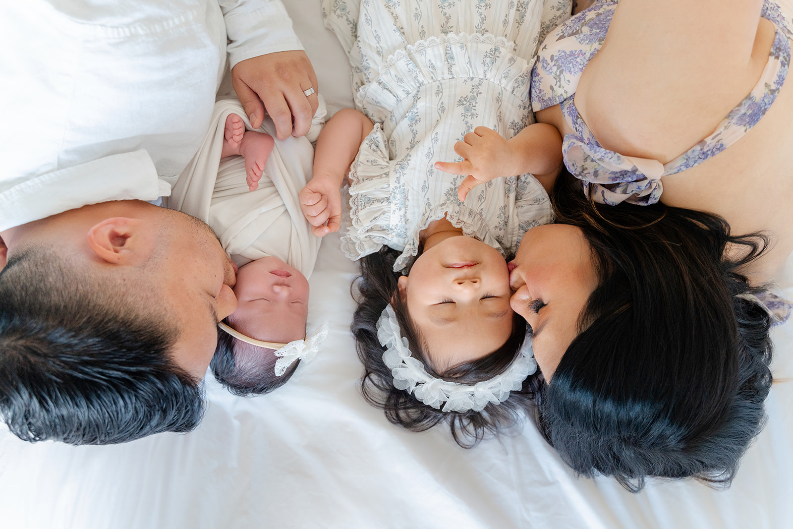 A mom and dad kiss their newborn and toddler daughters on the cheek while laying on a bed after visiting Community Roots Academy