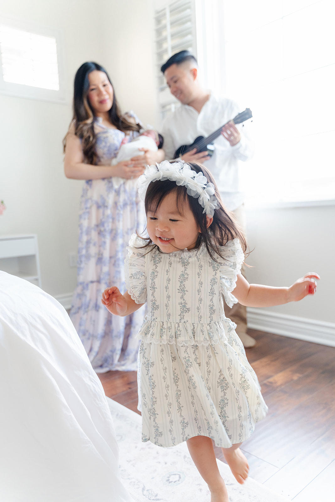 A toddler girl runs in a bedroom as mom and dad stand in the corner watching and playing ukelele for the newborn after finding Community Roots Academy