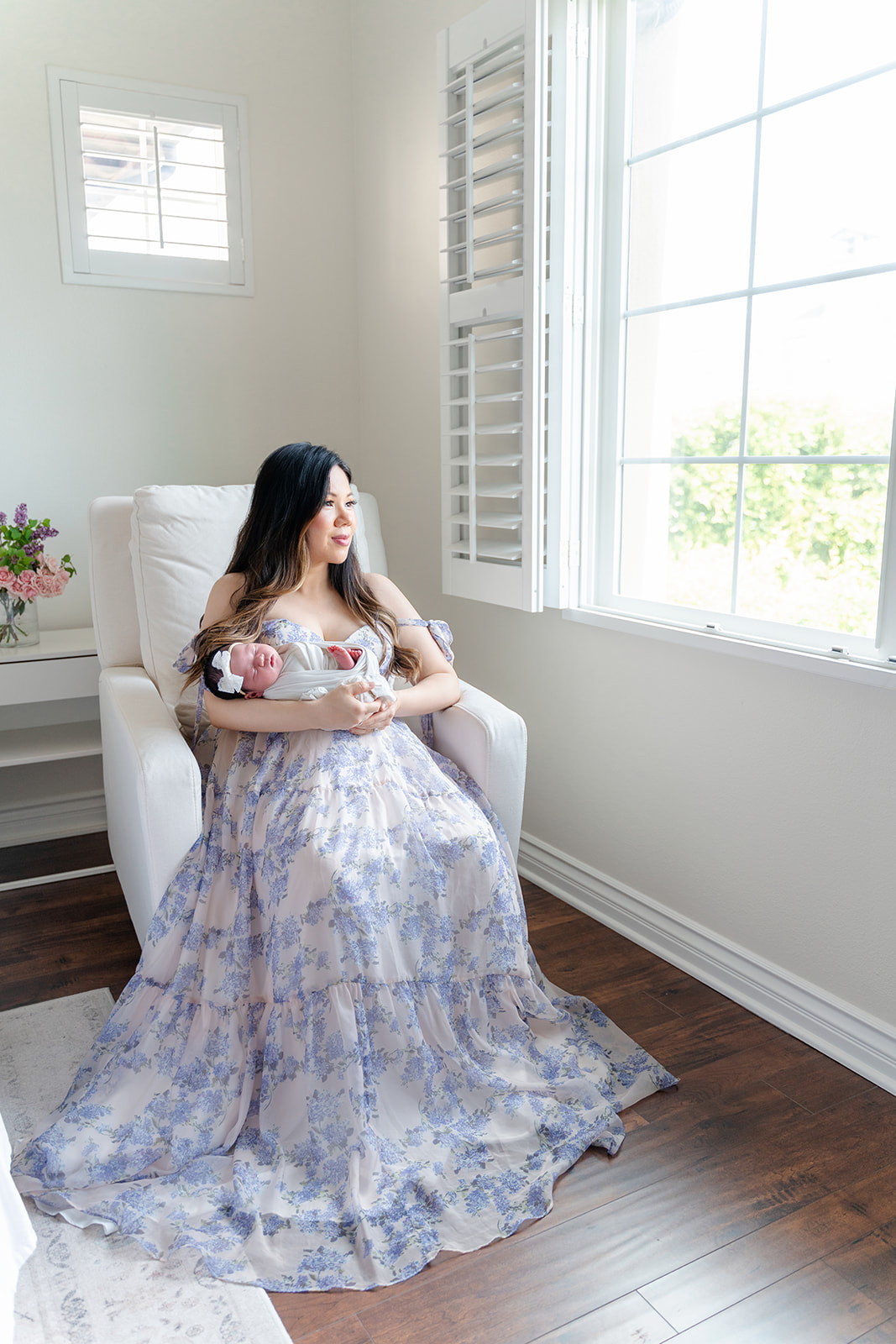 A new mother in a long blue maternity gown sits in a nursing chair in a window holding her sleeping newborn after visiting Community Roots Academy