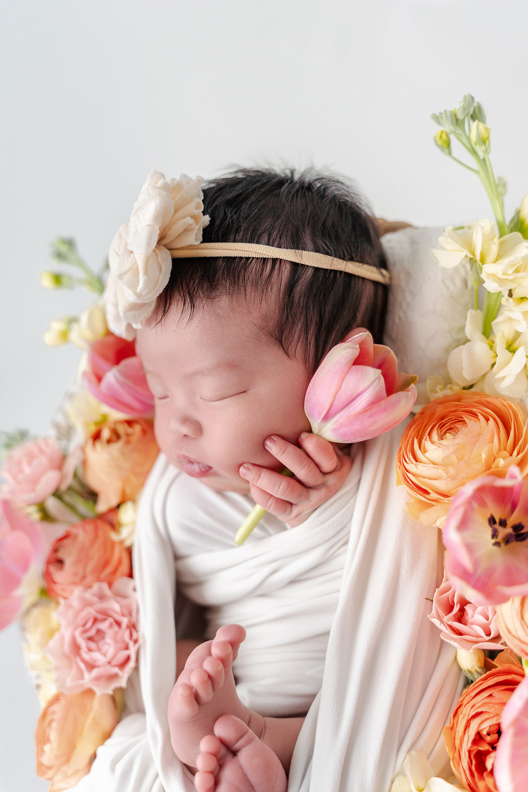 A closeup of a newborn baby sleeping surrounded by flowers in a white swaddle
