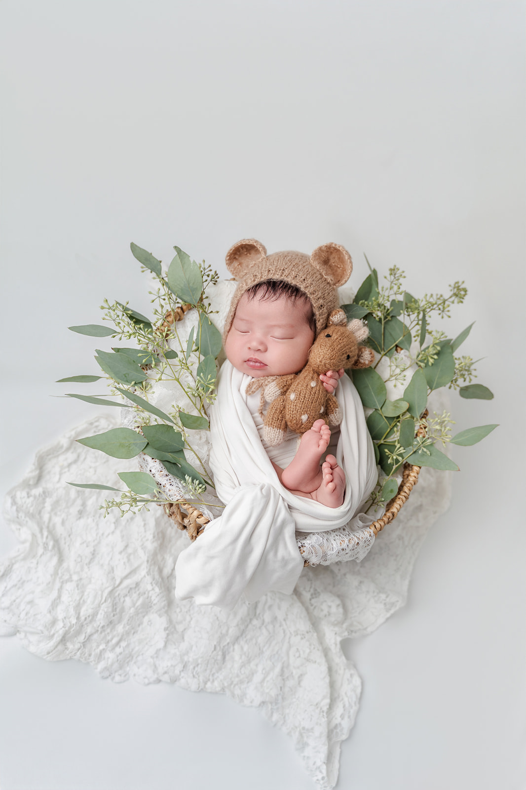 A newborn baby sleeps in a wicker basket holding a knit stuffed giraffe with white blankets and greenery