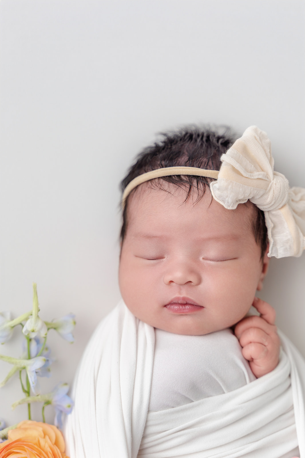 A closeup of a newborn baby girl sleeping in a white swaddle and cream bow next to flowers before visiting Northwood Montessori School