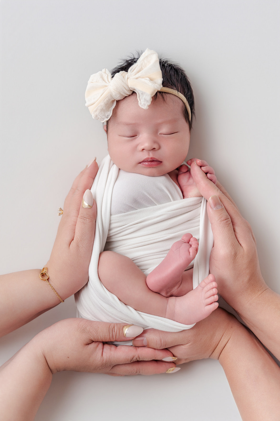 A newborn baby sleeps in a white swaddle and bow with mom and dad's hands on her before visiting Northwood Montessori School
