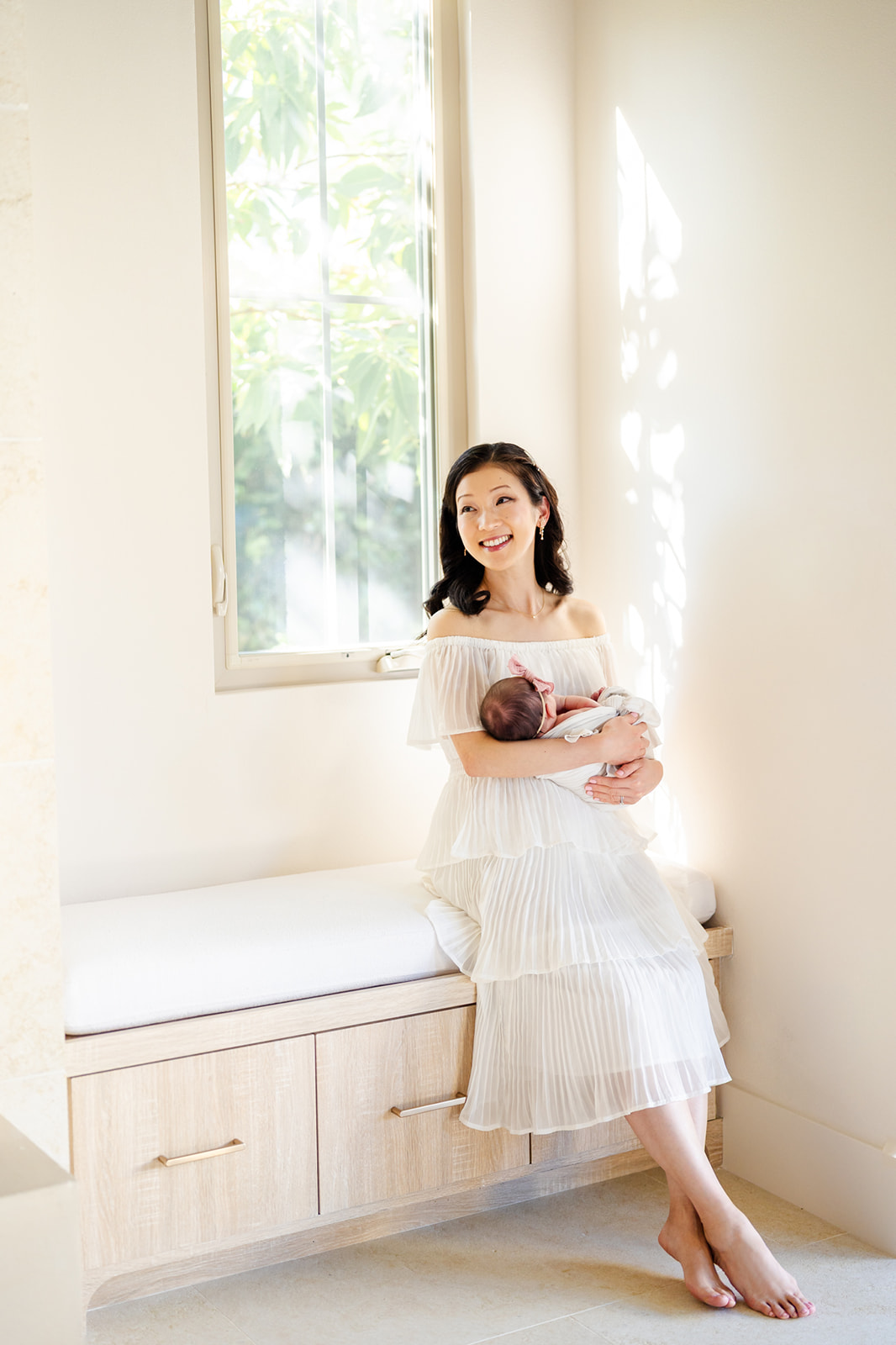 A smiling new mom in a white dress sits in a window bench cradling her sleeping newborn