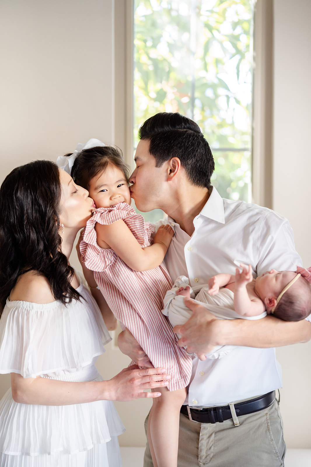 Mom and dad kiss their giggling toddler daughter as dad holds a newborn in his other arm