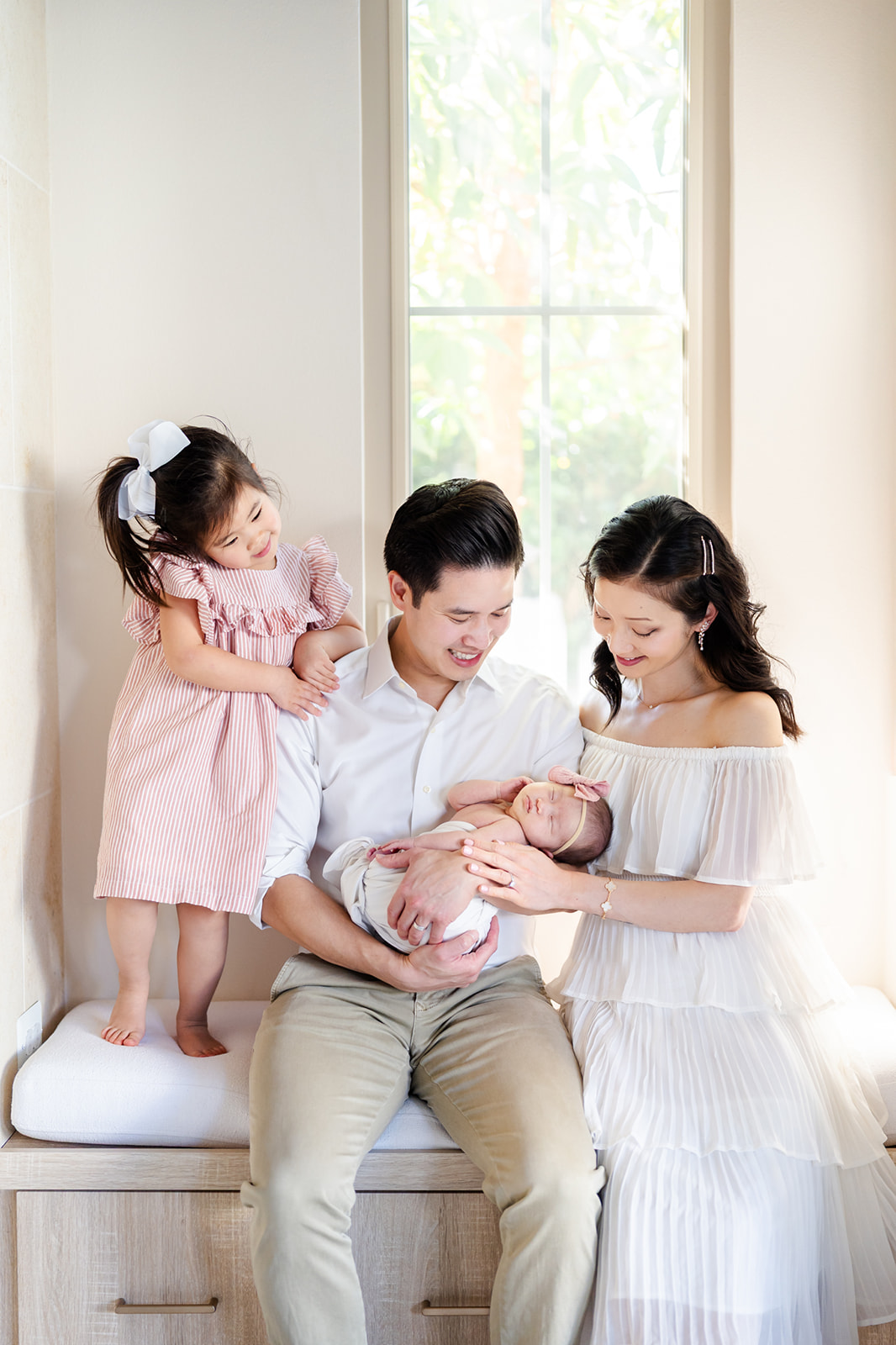 A dad sits in a window holding his sleeping newborn daughter as toddler girl leans on his shoulder and mom on the other side before visiting The Rinks Irvine