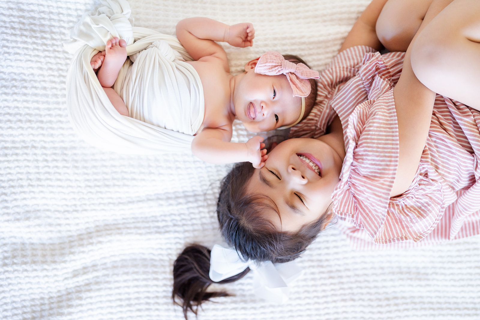 A giggling toddler girl lays on a bed in a red stripe dress head to head with her happy newborn sister before visiting The Rinks Irvine